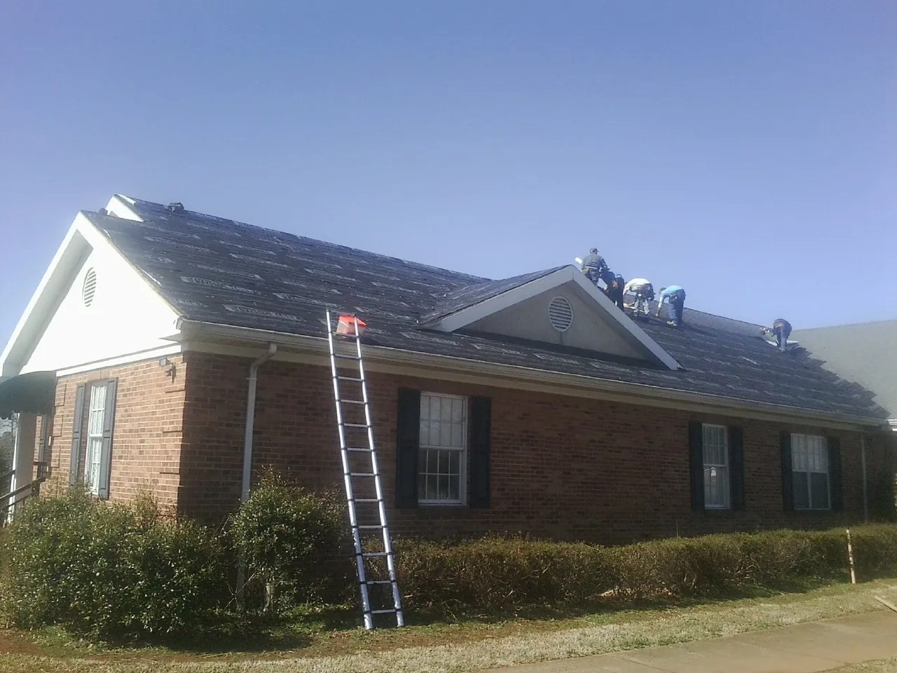 Crew tearing shingles off a brick ranch roof with a ladder set — Emergency Roof Repair work by our crew in Deale
